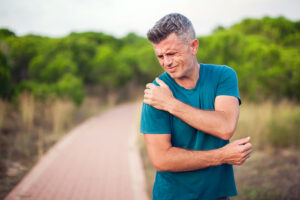 Athletic man wearing blue T-shirt on jogging path clutching his painful shoulder